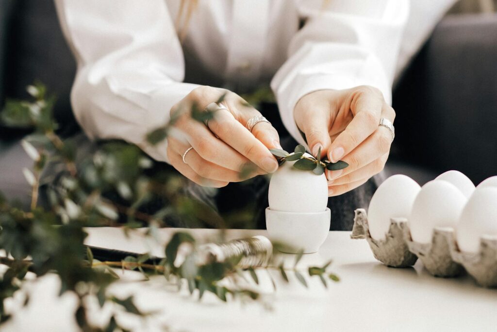 Photo by ROMAN ODINTSOV Hands arranging leaves on white eggs for an Easter decoration indoors, creating a natural festive look.
